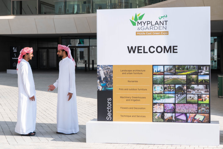 Two men in traditional Emirati attire speaking near the large welcome sign at Myplant & Garden Middle East.