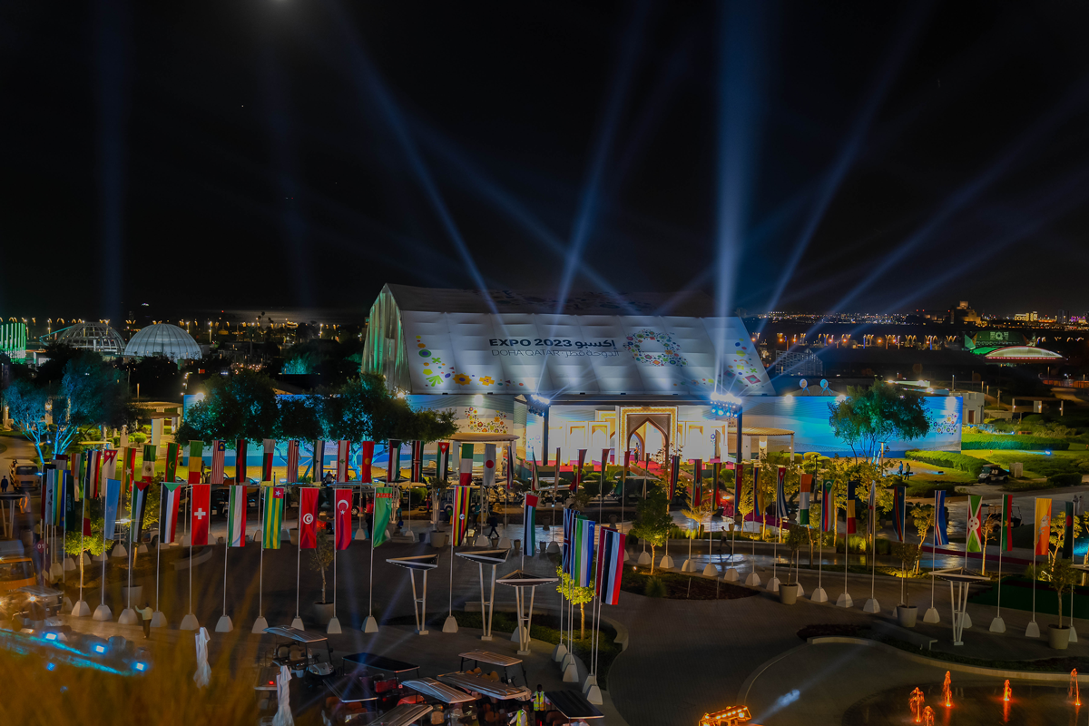 Night view of Expo 2023 Doha site with illuminated pavilions and rows of international flags.