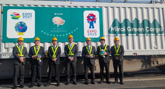 Group of officials in safety helmets stand beside a freight train container branded for Expo 2025 and GREEN×EXPO 2027 tree transport project