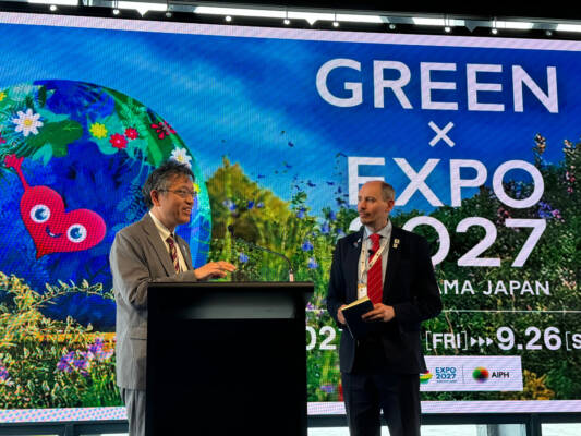 Speaker at a podium in front of a GREEN×EXPO 2027 Yokohama presentation screen during an AIPH meeting.