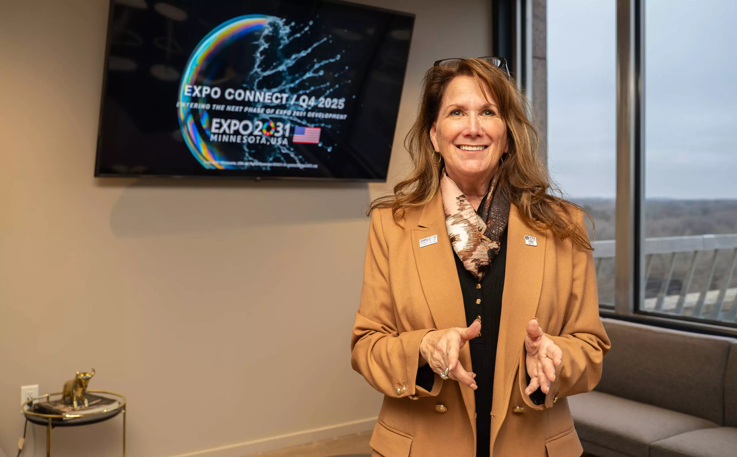 Wendy Meadley stands in an office with a screen behind her displaying the Expo 2031 Minnesota branding.